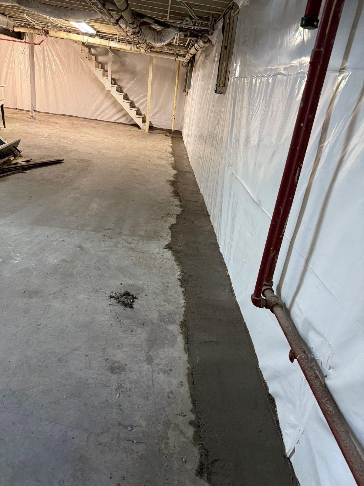 Completed interior basement drain with freshly re-poured concrete perimeter against a white vapor-barrier wall